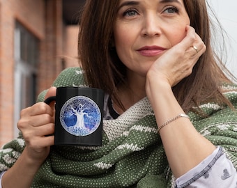 May include: Black ceramic mug featuring a detailed tree design within a circular frame. The tree has a white trunk and branches, set against a blue and purple background. The mug is held by a person wearing a green shawl.