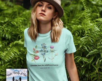 Könnte beinhalten: Hellblaues T-Shirt mit einem Aquarell-Blumenmuster und dem Text "Betrachtet die Lilien auf dem Felde" und "Lukas 12,25-28". Das Shirt hat auch ein kleines Logo unten links mit "Faith, Family & Fun" und "TrueFoundationDesign".