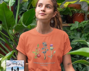 Könnte beinhalten: Orangefarbenes T-Shirt mit einem Aquarell-Blumenmuster und dem Text "Seht, wie die Lilien wachsen" aus Lukas 12,25-28. Das T-Shirt enthält auch das Logo von True Foundation Design.