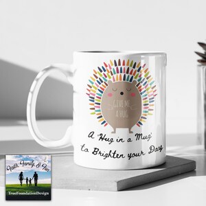 May include: White ceramic mug featuring a cartoon hedgehog with colorful quills and the text "GIVE ME A HUG". The mug also has the text "A 'Hug in a Mug' to Brighten your Day". The mug is a heartwarming gift.
