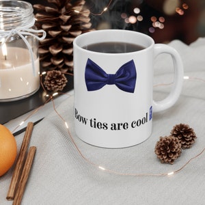 May include: White ceramic mug featuring a navy blue bow tie graphic and the text "Bow ties are cool". The mug is filled with a dark liquid and has a white handle. The mug is on a light gray surface.
