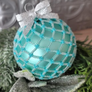 May include: A spherical Christmas ornament with a matte light blue finish, encased in a decorative lattice of silver and bright blue beads. A small, shimmering silver ribbon bow is attached to the top, serving as a hanging loop for the ornament.