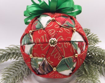 May include: A red, green, and gold fabric Christmas ornament with a green bow on top. The ornament is decorated with gold trim and beads. The fabric has a pattern of holly leaves, berries, and the words "Merry Christmas".
