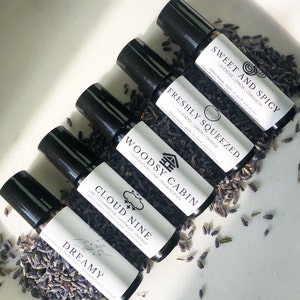 May include: Six black roller bottles with white labels and different scents. The labels have names like "Sweet and Spicy", "Freshly Squeezed", "Woodsy Cabin", "Cloud Nine", and "Dreamy". The bottles are arranged in a row on a white surface with dried lavender flowers scattered around them.