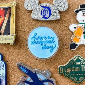 May include: A blue and white round pin with glitter and the text "Have a magical day" in white cursive lettering.