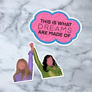 May include: Two cartoon figures, one with brown hair and a purple shirt and the other with black hair and a green shirt, are holding hands and jumping in the air. A pink speech bubble with blue trim says "This is what dreams are made of."