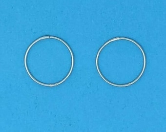May include: Two small, silver-colored hoop earrings are displayed against a bright blue background. The earrings are circular and appear to be made of a thin metal wire. The earrings are simple and minimalist in design.