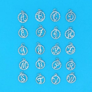 Op de afbeelding: Een set van 26 zilveren bedels, elk met een andere letter van het alfabet in een cursief lettertype binnen een cirkelvormig frame. De bedels zijn in alfabetische volgorde gerangschikt op een blauwe achtergrond.