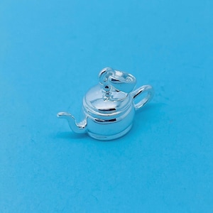 May include: Silver charm shaped like a teapot with a shiny finish. The teapot has a spout, a handle and a lid.