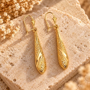 Puede incluir: Un par de pendientes colgantes de oro con forma de lágrima alargada y complejos patrones de volutas grabados. Los pendientes cuelgan de ganchos de alambre curvados, mostrando un acabado metálico pulido y texturas detalladas.
