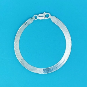 Könnte beinhalten: Ein silbernes Fischgrätenarmband mit sicherem Verschluss, vor einem leuchtend blauen Hintergrund. Das flache, verbundene Design des Armbands reflektiert das Licht und zeigt seine polierte Oberfläche. Ein klassisches Schmuckstück.