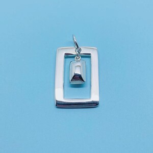 May include: A silver pendant featuring a square frame with a smaller square shape suspended within. The pendant is displayed against a light blue backdrop.