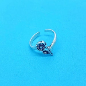 Könnte beinhalten: Ein silberfarbener, verstellbarer Ring mit floralem Design. Der Ring hat eine detaillierte Blume mit mehreren Blütenblättern und einem kleinen Blatt. Der Ring ist vor einem hellblauen Hintergrund platziert.
