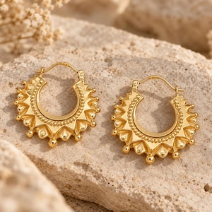 May include: A pair of gold hoop earrings featuring a sunburst design with a beaded inner rim and pointed triangular outer edges. Each earring has a hinged post closure and a polished metallic finish, showcasing a detailed radial pattern.
