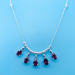Può includere: Una collana d'argento con una fila di piccole perle e cinque gocce di pietre preziose rosse pendenti.
