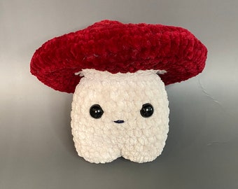 May include: A crocheted plush toy shaped like a mushroom. The mushroom has a red cap and a white body with black eyes and a small mouth.