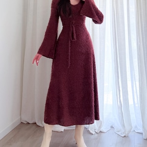 May include: A long, burgundy dress with a flared skirt and long sleeves. The dress features a tie belt at the waist and is paired with cream-colored ankle boots. The fabric appears to be a soft, textured material.