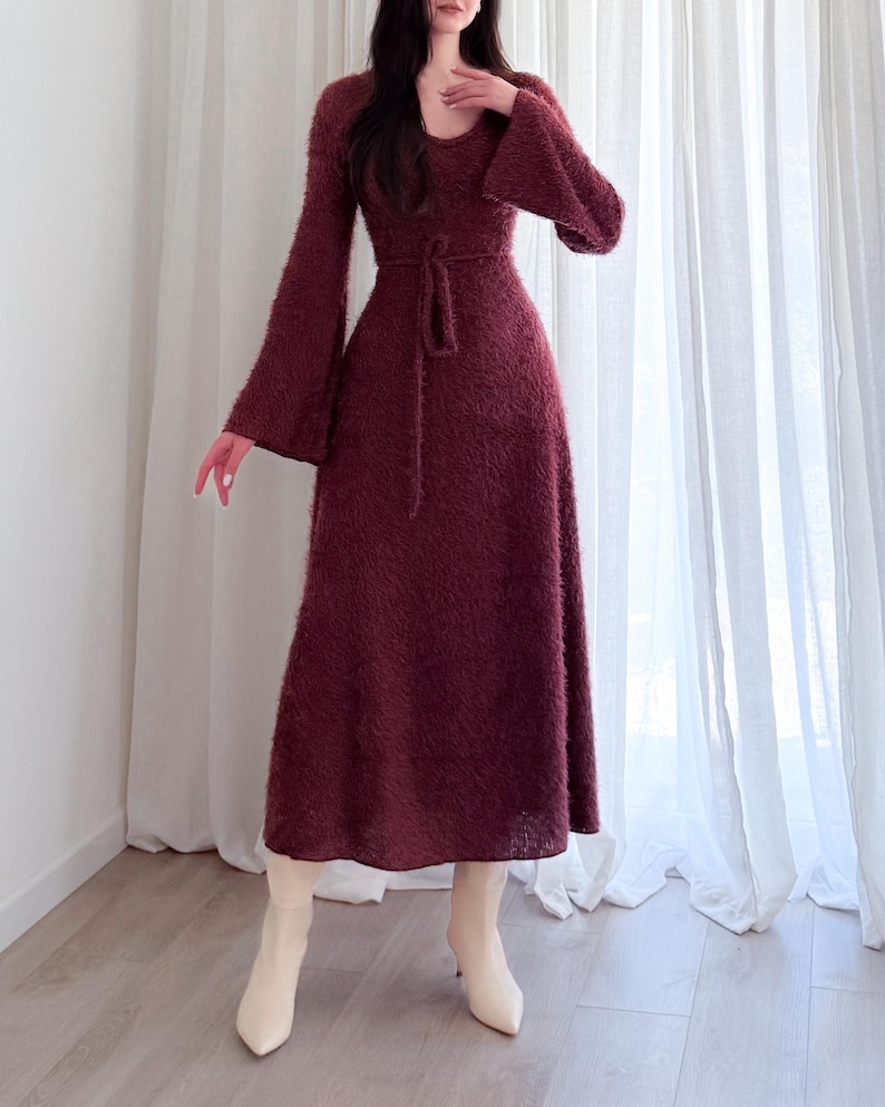 May include: A long, burgundy dress with a flared skirt and long sleeves. The dress features a tie belt at the waist and is paired with cream-colored ankle boots. The fabric appears to be a soft, textured material.