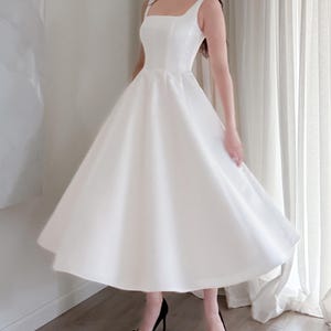 May include: A white, sleeveless dress with a square neckline and a flared skirt. The dress is paired with black high-heeled shoes. The dress appears to be made of a smooth, possibly satin-like fabric, and is knee-length.