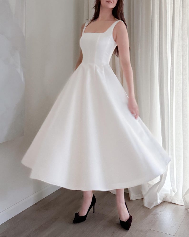 May include: A white, sleeveless dress with a square neckline and a flared skirt. The dress is paired with black high-heeled shoes. The dress appears to be made of a smooth, possibly satin-like fabric, and is knee-length.