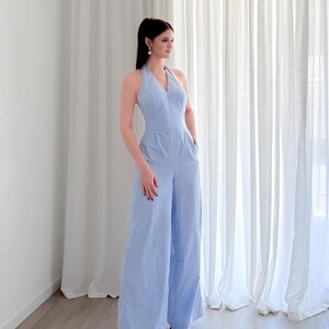 May include: A light blue halter-neck jumpsuit with wide legs. The jumpsuit has a V-neckline and pockets. The person is wearing white sandals. The background features white curtains and a light-colored wooden floor.