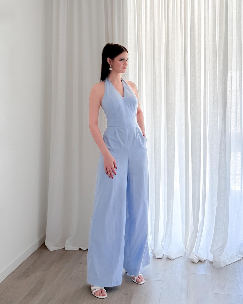 May include: A light blue halter-neck jumpsuit with wide legs. The jumpsuit has a V-neckline and pockets. The person is wearing white sandals. The background features white curtains and a light-colored wooden floor.
