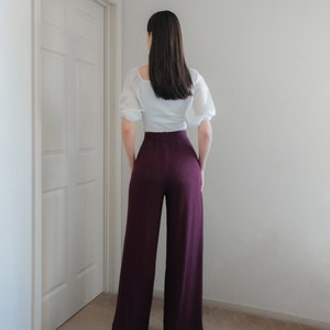 Op de afbeelding: Een persoon draagt een witte blouse met korte, pofmouwen en een hoog getailleerde, wijd uitlopende, bordeauxrode broek. De outfit is eenvoudig en elegant, geschikt voor diverse gelegenheden. De broek heeft een getextureerde uitstraling.