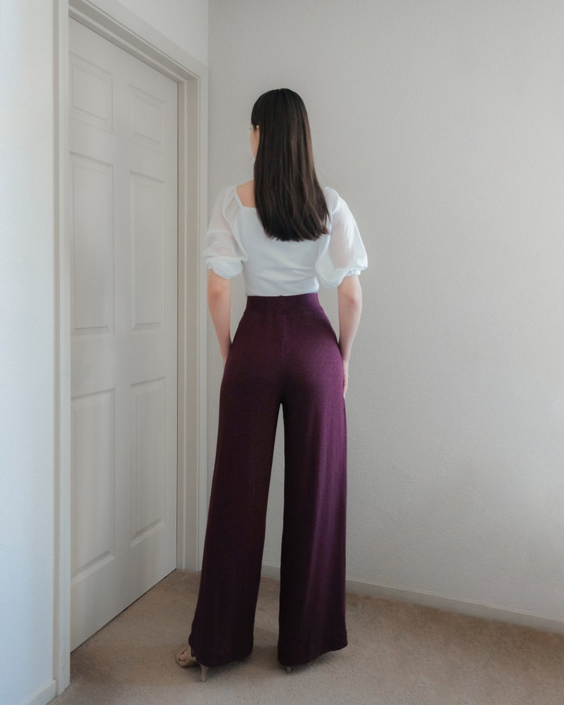 Op de afbeelding: Een persoon draagt een witte blouse met korte, pofmouwen en een hoog getailleerde, wijd uitlopende, bordeauxrode broek. De outfit is eenvoudig en elegant, geschikt voor diverse gelegenheden. De broek heeft een getextureerde uitstraling.