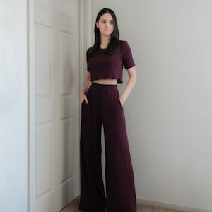 Op de afbeelding: Een bordeauxrode tweedelige outfit bestaande uit een crop top met korte mouwen en een broek met wijde pijpen. De broek heeft zakken. De outfit is gemaakt van een zachte, getextureerde stof. De persoon draagt beige sandalen.