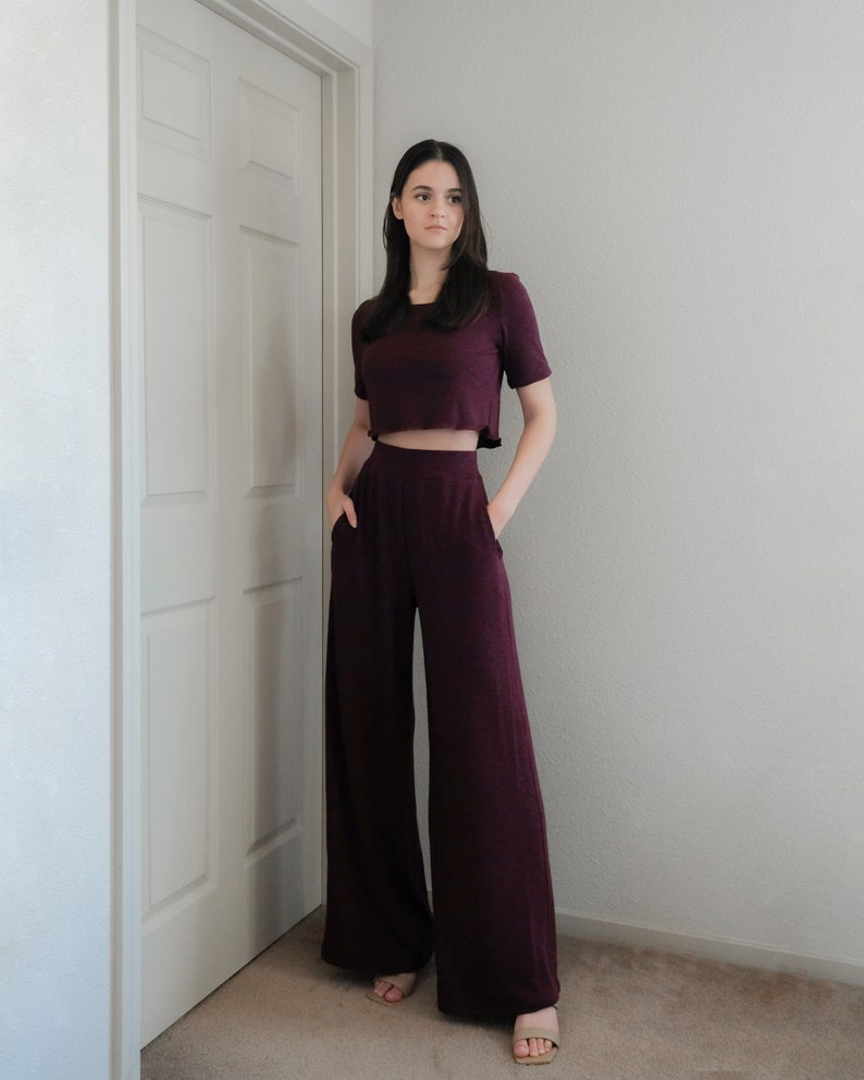 Op de afbeelding: Een bordeauxrode tweedelige outfit bestaande uit een crop top met korte mouwen en een broek met wijde pijpen. De broek heeft zakken. De outfit is gemaakt van een zachte, getextureerde stof. De persoon draagt beige sandalen.