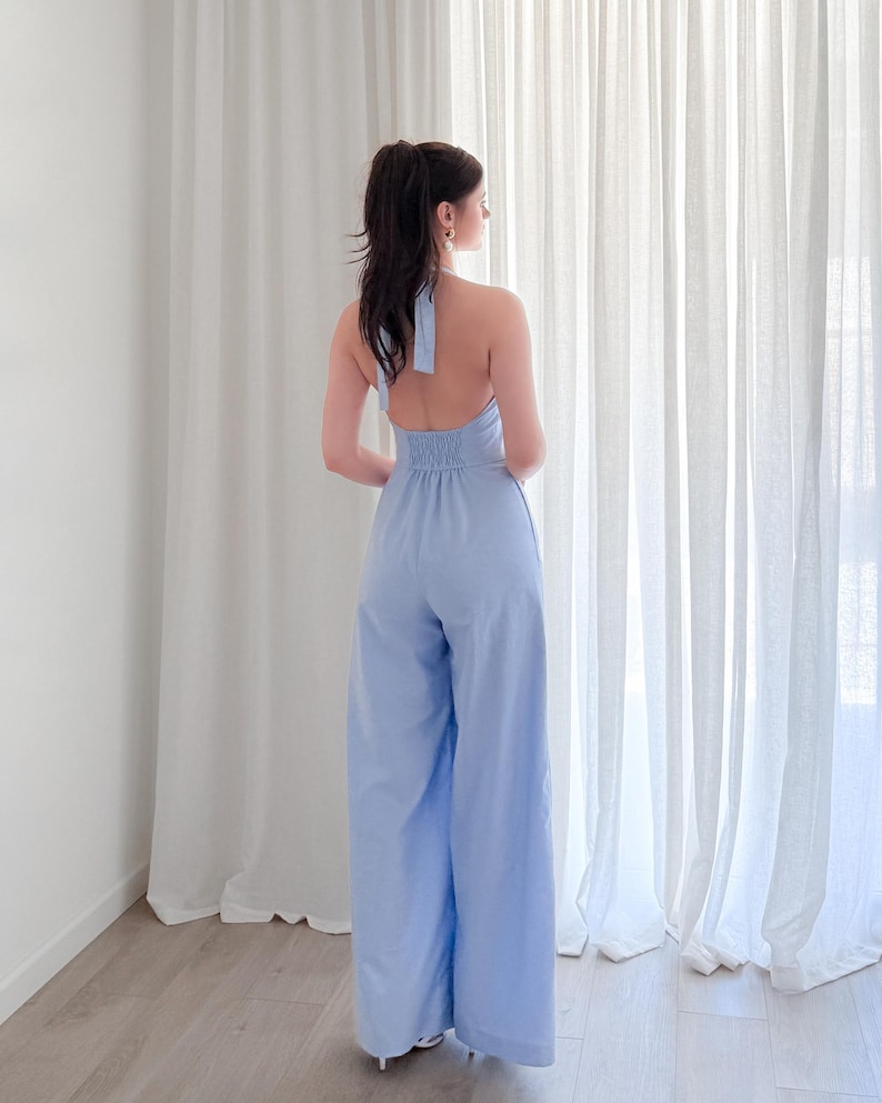 May include: A light blue halter jumpsuit with wide legs. The jumpsuit features a halter neckline and a gathered waist. The model is standing in front of a white curtain.