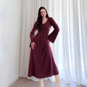 May include: A burgundy-colored, long-sleeved dress with a V-neck and flared sleeves. The dress has a tie belt at the waist and a midi-length skirt. The person is wearing cream-colored boots.