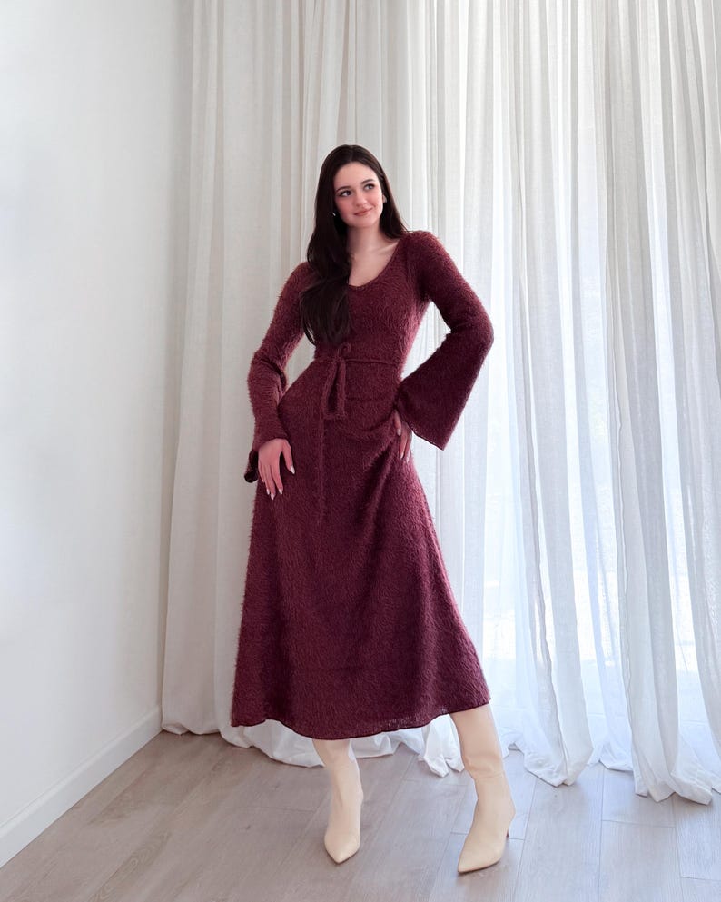 May include: A burgundy-colored, long-sleeved dress with a V-neck and flared sleeves. The dress has a tie belt at the waist and a midi-length skirt. The person is wearing cream-colored boots.