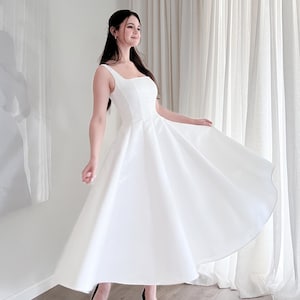 May include: A white midi dress with a square neckline and a flared skirt. The dress is paired with black heels. The dress is made of a smooth, opaque fabric and appears to be a formal or special occasion garment.