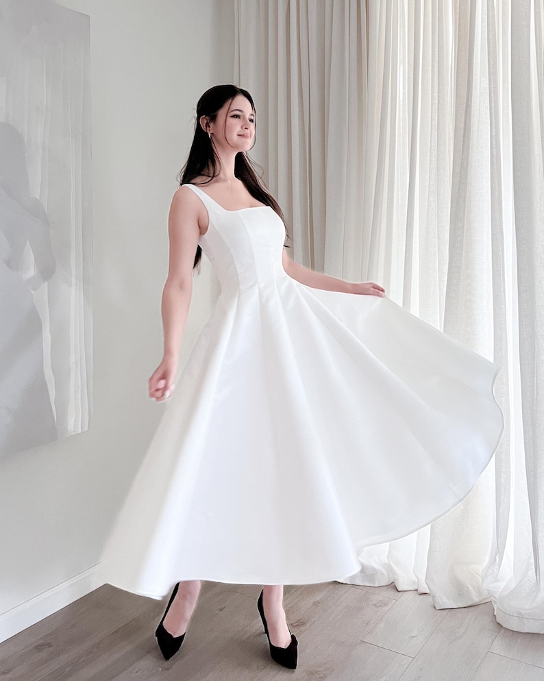 May include: A white midi dress with a square neckline and a flared skirt. The dress is paired with black heels. The dress is made of a smooth, opaque fabric and appears to be a formal or special occasion garment.