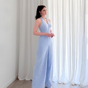 May include: A light blue halter jumpsuit with wide legs. The jumpsuit has a V-neck and is paired with white sandals. The model is standing in front of a white curtain.