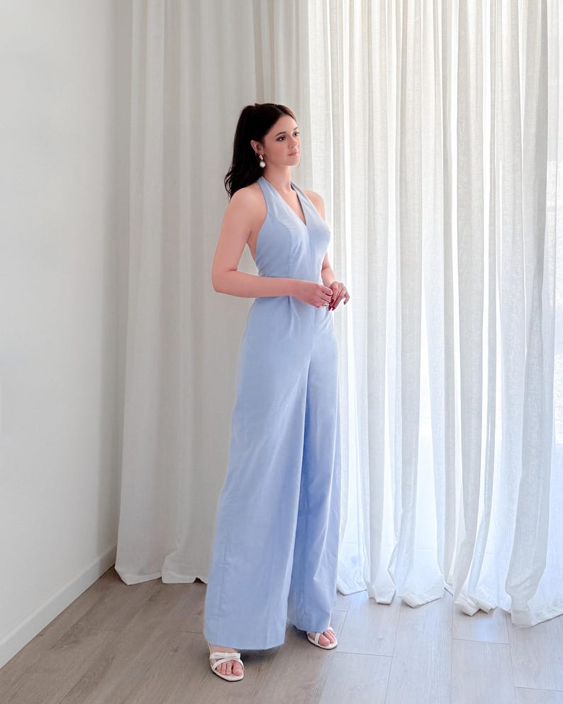 May include: A light blue halter jumpsuit with wide legs. The jumpsuit has a V-neck and is paired with white sandals. The model is standing in front of a white curtain.