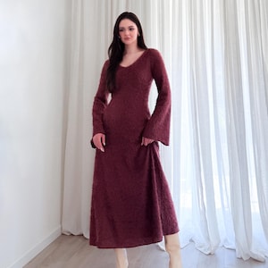 May include: A burgundy-colored, long-sleeved dress with a V-neck and flared sleeves. The dress has a textured, fuzzy appearance and falls to the mid-calf. The model is wearing cream-colored boots.