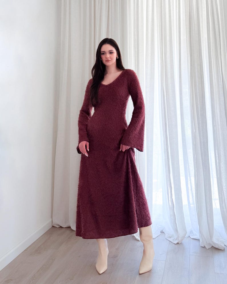 May include: A burgundy-colored, long-sleeved dress with a V-neck and flared sleeves. The dress has a textured, fuzzy appearance and falls to the mid-calf. The model is wearing cream-colored boots.