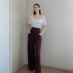 Op de afbeelding: Een witte top met een vierkante halslijn en korte, doorschijnende mouwen, gecombineerd met een wijde, bordeauxrode broek. De broek heeft zakken. De outfit wordt afgemaakt met beige sandalen. Het model staat bij een witte deur en muur.