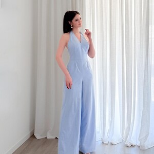 May include: A light blue halter jumpsuit with wide legs. The jumpsuit has a V-neck and a fitted waist. The model is wearing white sandals. The background is a white wall and white curtains.