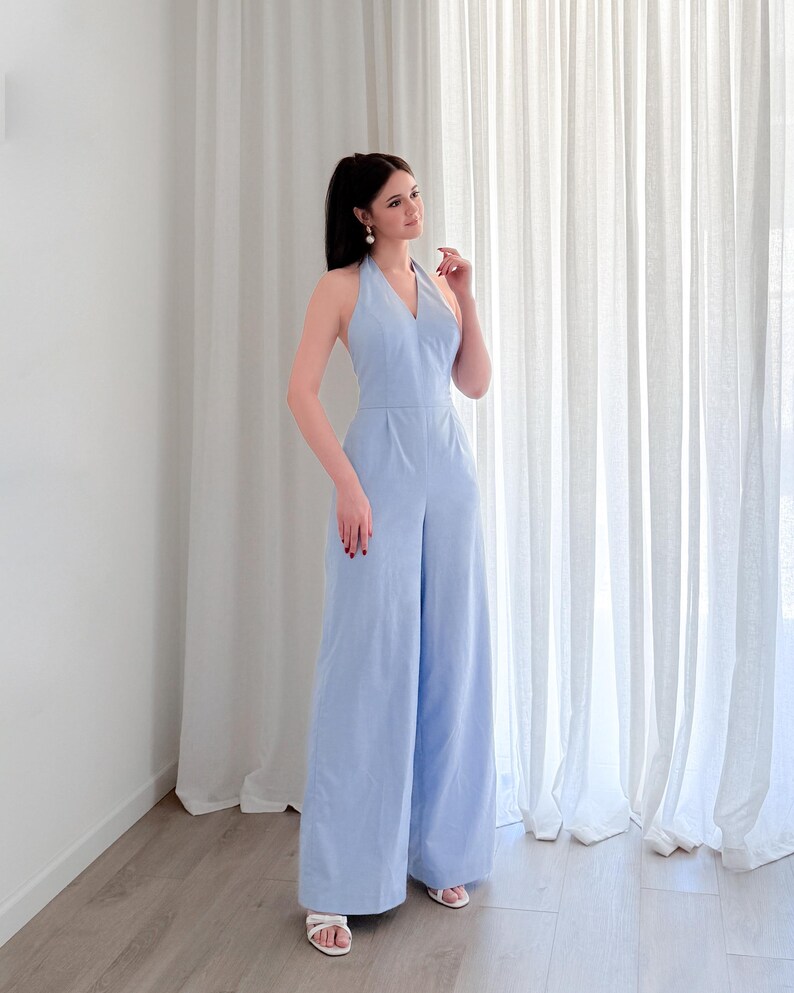 May include: A light blue halter jumpsuit with wide legs. The jumpsuit has a V-neck and a fitted waist. The model is wearing white sandals. The background is a white wall and white curtains.