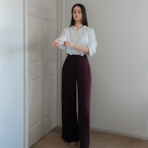 Op de afbeelding: Een vrouw draagt een wit overhemd en een wijde, donker bordeauxrode broek. De broek met hoge taille is gemaakt van een zachte stof. De outfit wordt gecompleteerd met sandalen in neutrale kleuren.