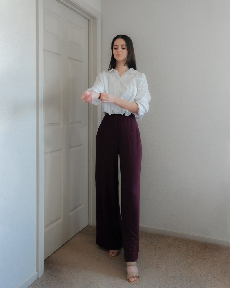 Op de afbeelding: Een vrouw draagt een wit overhemd en een wijde, donker bordeauxrode broek. De broek met hoge taille is gemaakt van een zachte stof. De outfit wordt gecompleteerd met sandalen in neutrale kleuren.
