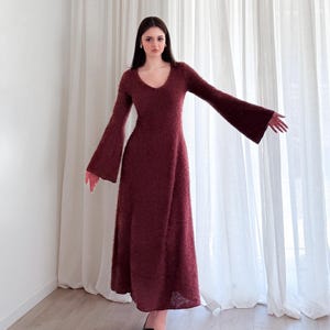 May include: A burgundy-colored, long-sleeved dress with a V-neck and flared sleeves. The dress has a textured appearance and falls to the ankles. The model is wearing black pointed-toe heels. The background features white curtains and a light-colored floor.
