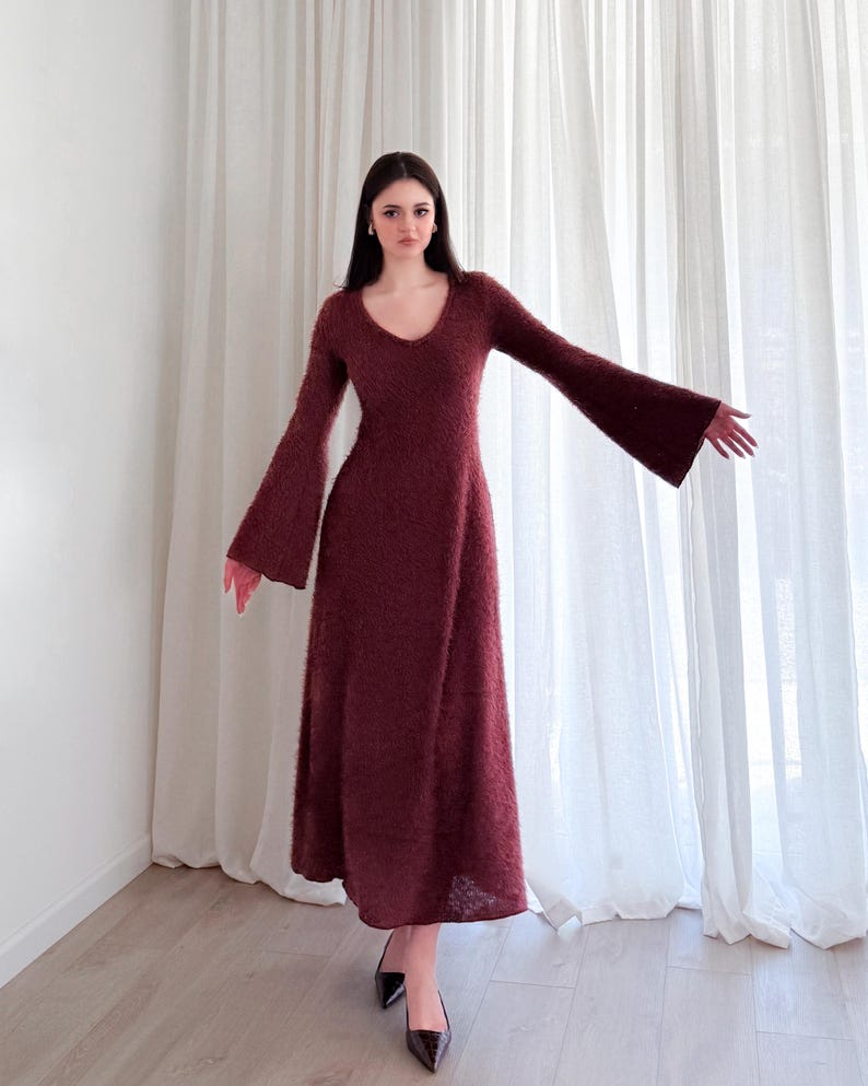 May include: A burgundy-colored, long-sleeved dress with a V-neck and flared sleeves. The dress has a textured appearance and falls to the ankles. The model is wearing black pointed-toe heels. The background features white curtains and a light-colored floor.