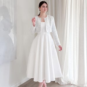 May include: A white midi dress with a square neckline is paired with a light blue tweed jacket. The jacket has long sleeves and a cropped fit. The dress has a full skirt. The person is wearing silver heels.