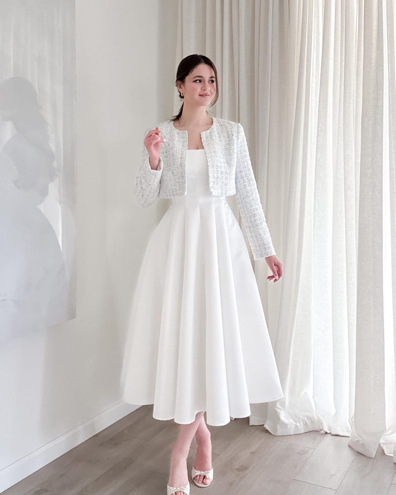 May include: A white midi dress with a square neckline is paired with a light blue tweed jacket. The jacket has long sleeves and a cropped fit. The dress has a full skirt. The person is wearing silver heels.