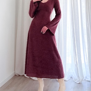 May include: A long, burgundy-colored dress with a V-neck and flared sleeves. The dress has a fuzzy texture and is paired with cream-colored ankle boots. The dress is floor-length and appears to be made of a soft material.