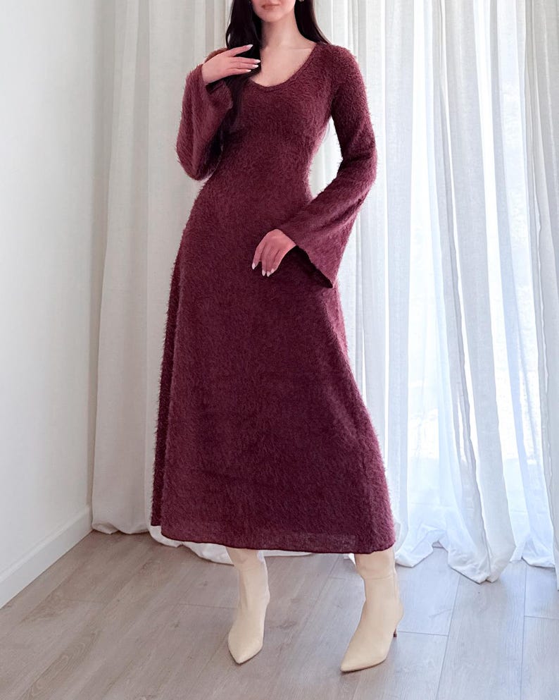May include: A long, burgundy-colored dress with a V-neck and flared sleeves. The dress has a fuzzy texture and is paired with cream-colored ankle boots. The dress is floor-length and appears to be made of a soft material.